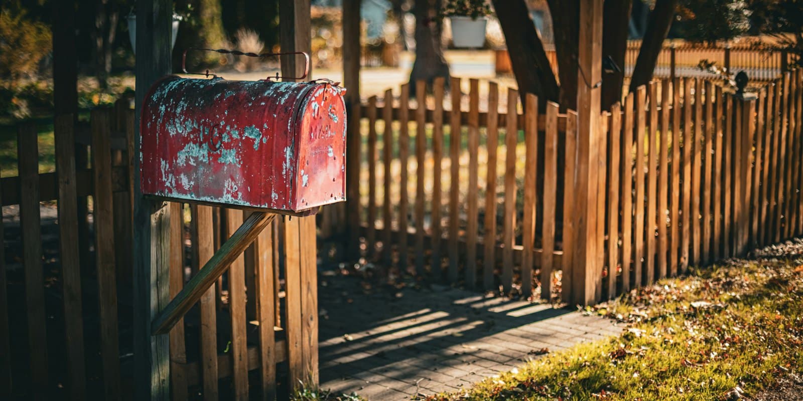 A weathered red mailbox on a post beside a wooden fence